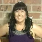 Karin Riebe standing in front of a brick fireplace, smiling and dressed in a purple gown.