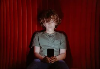 Child sitting alone looking at a smartphone, illuminated by screen light against a dark red background