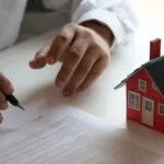Person reviewing and signing a contract next to a model red house representing a real estate transaction or consumer dispute