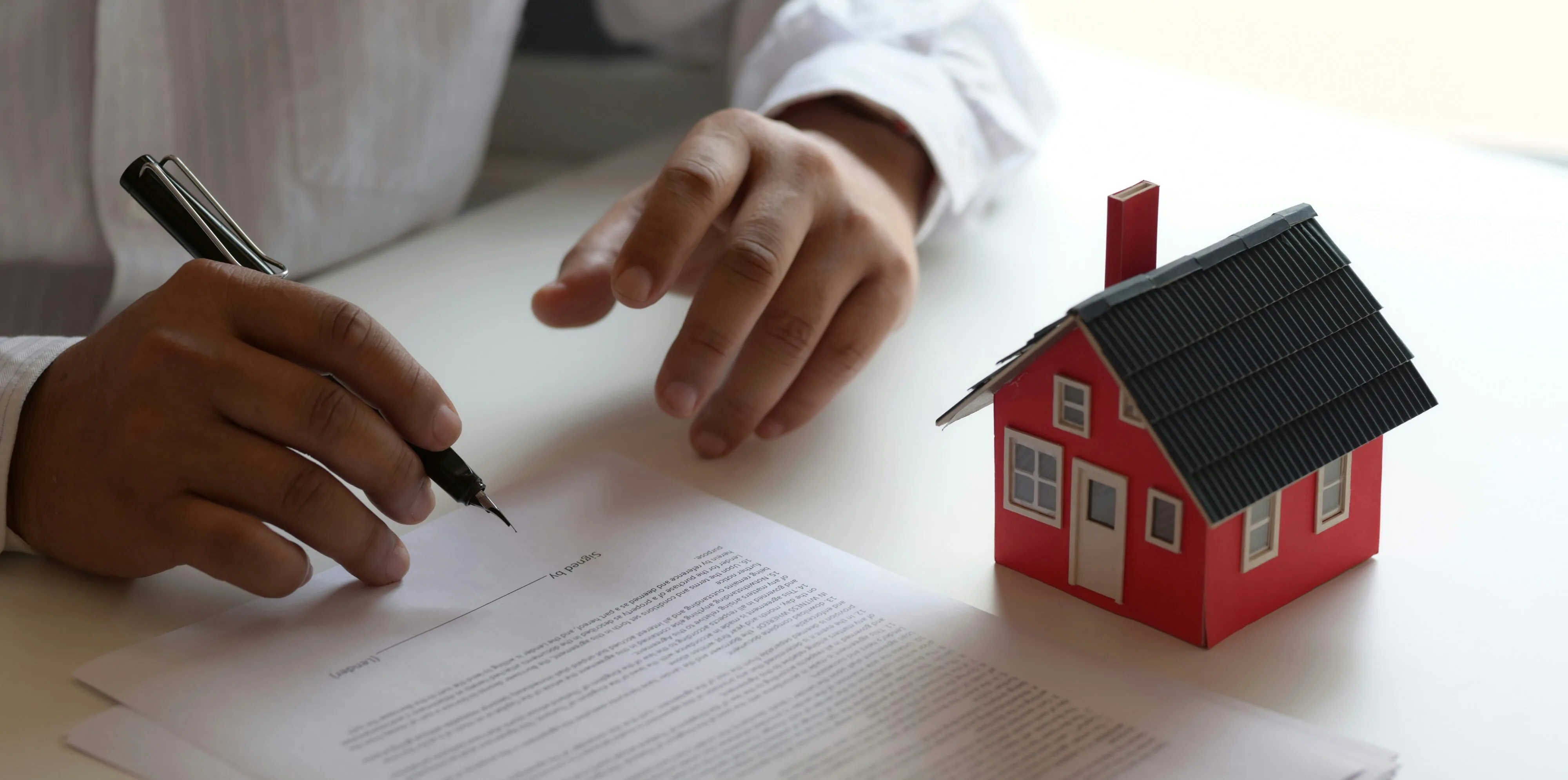 Person reviewing and signing a contract next to a model red house representing a real estate transaction or consumer dispute