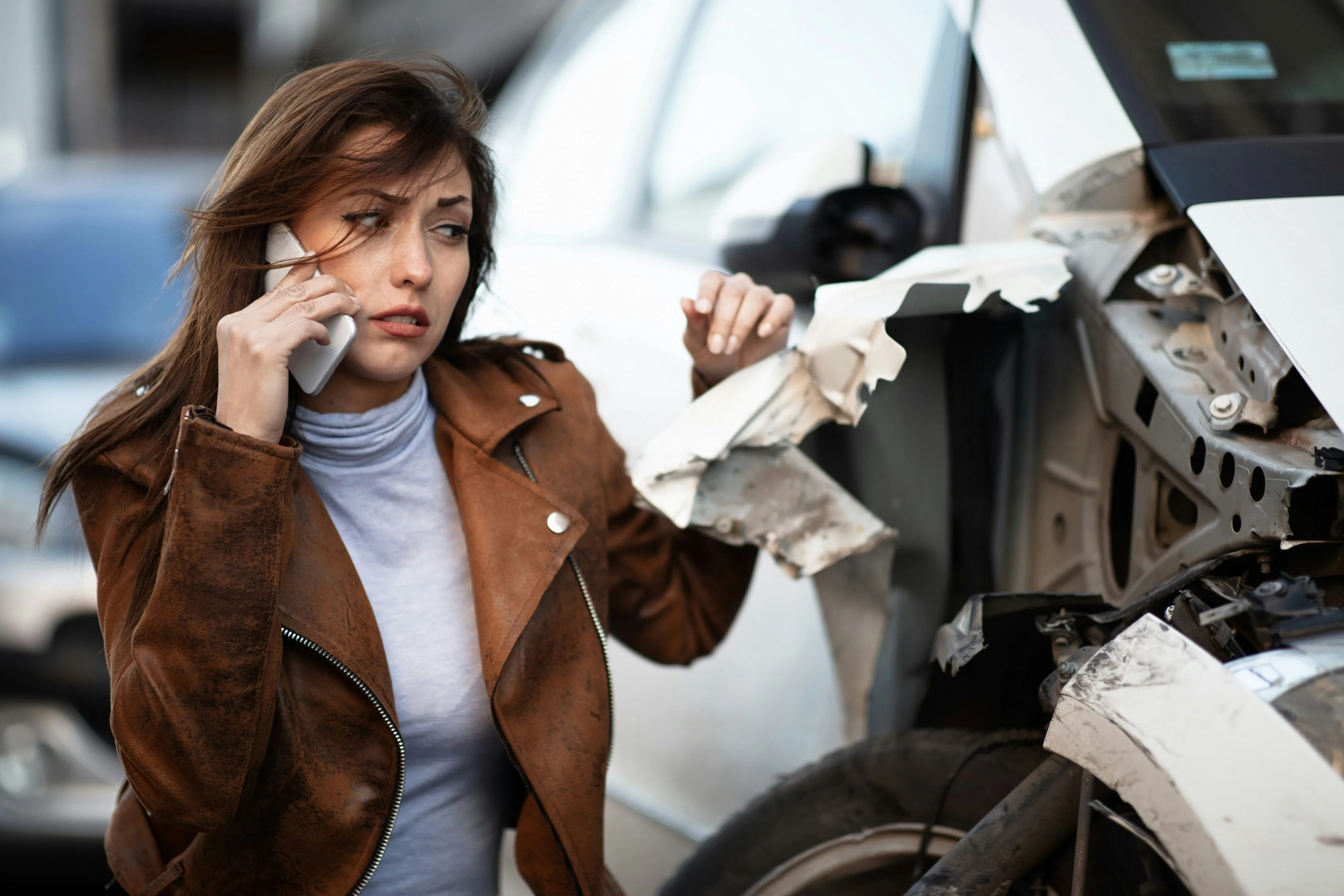 Driver standing beside a damaged vehicle after a collision while speaking on the phone with her insurance company.